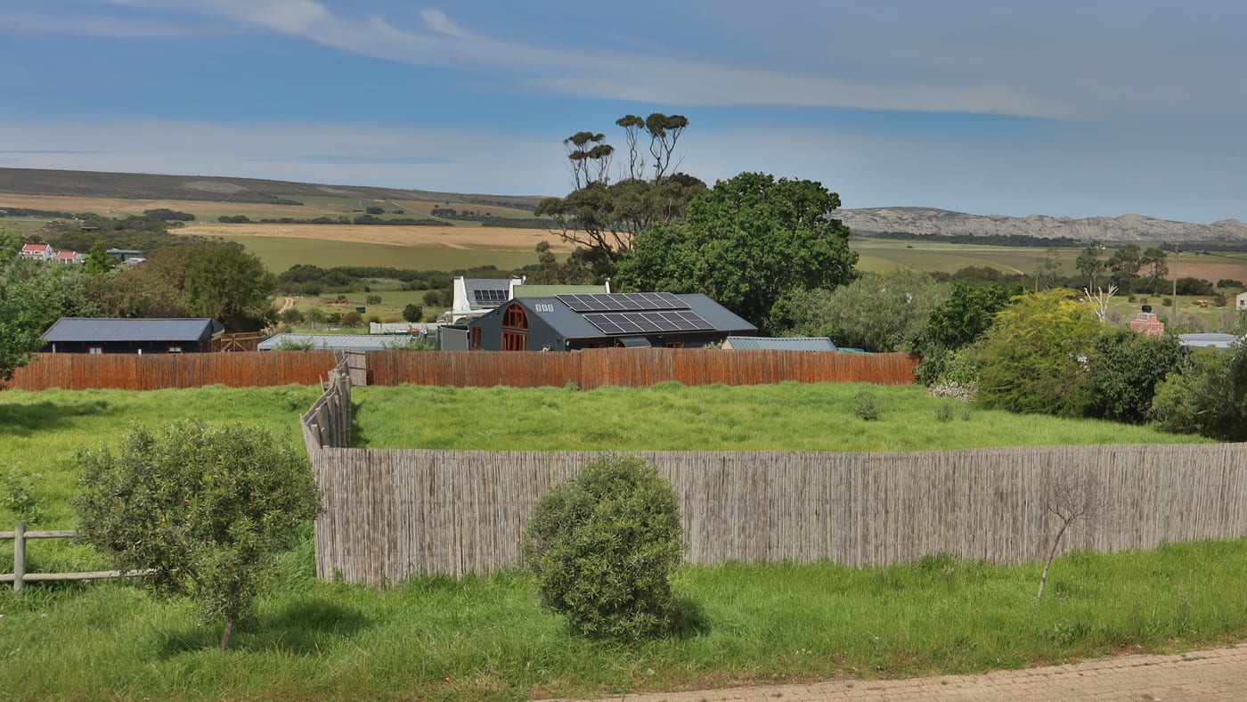 The stand inside its reed fences, seen from across the street, with village roofs, farmland and the hills beyond