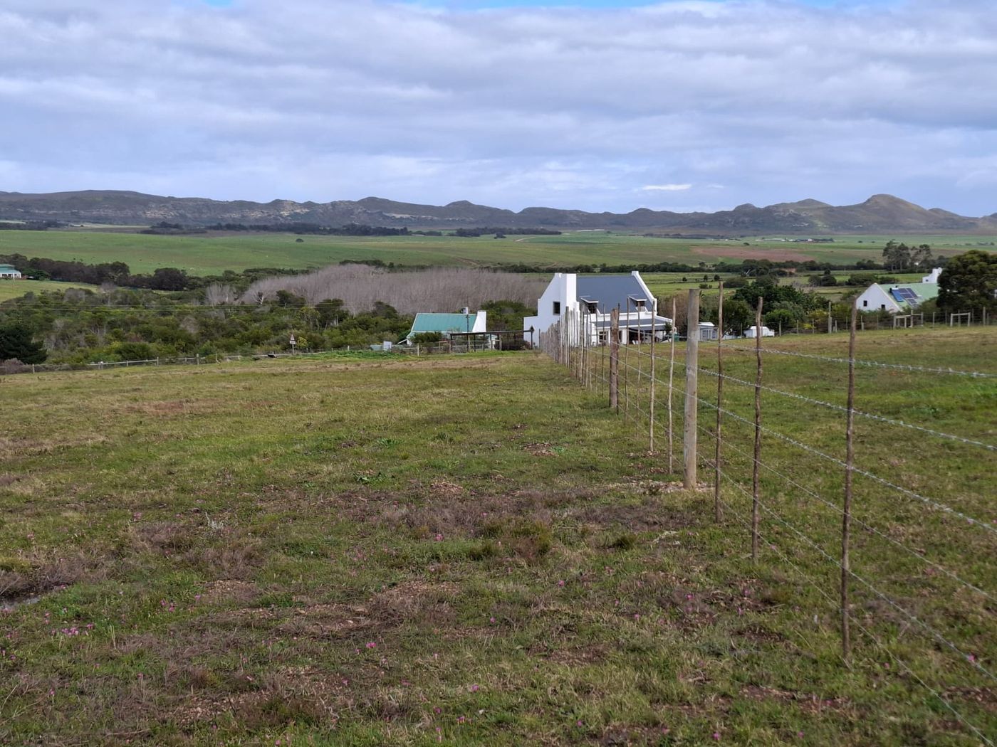 Ein Drahtzaun, der die Wiese hinabläuft, dahinter Häuser des Ortes, Farmland und die Berge