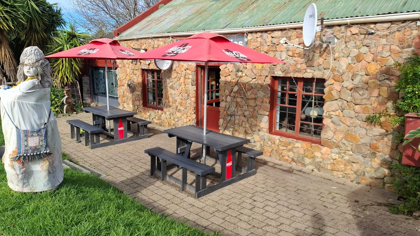 Das Pubgebäude aus Stein in Baardskeerdersbos, rote Sonnenschirme über den Tischen auf der gepflasterten Terrasse