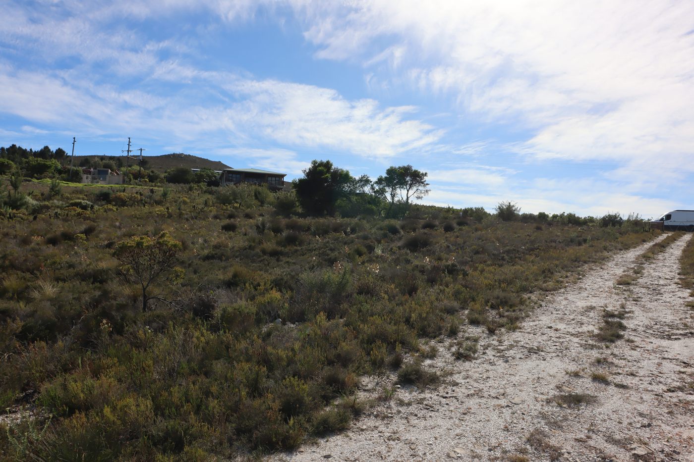 The sand road running up the slope beside the fynbos to a van parked at a fence, with houses on the hill to the left