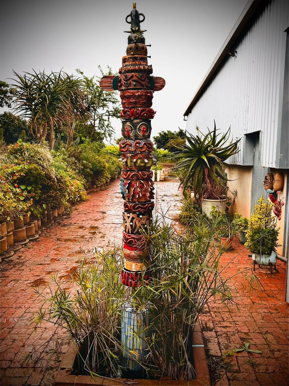 A brick path beside a building, a carved pole in a bed of grasses and aloes