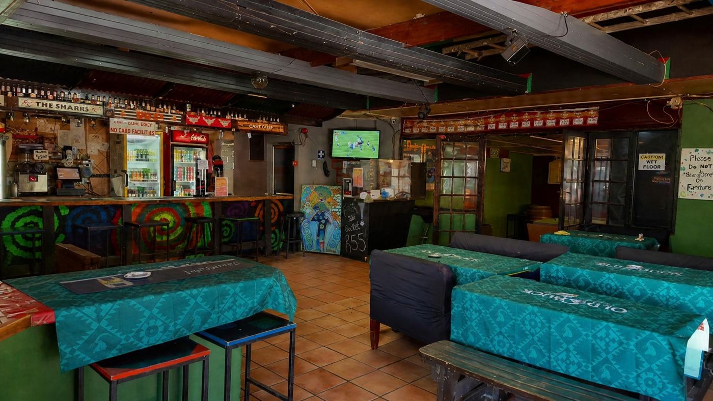 Die Hauptbar mit ihren Sitzplätzen: eine bemalte Theke mit Barhockern, Kühlschränke und ein Fernseher dahinter, Tische unter grünen Tischdecken