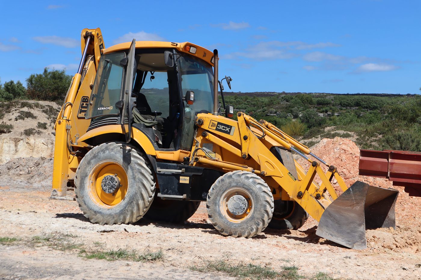 A yellow backhoe loader on sandy ground, with fynbos behind