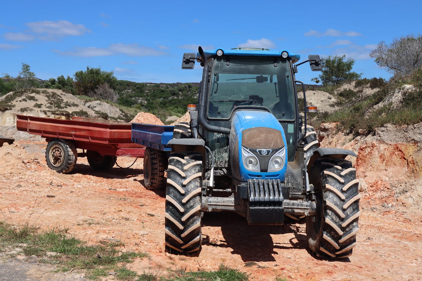 A blue tractor with a red trailer on sandy ground