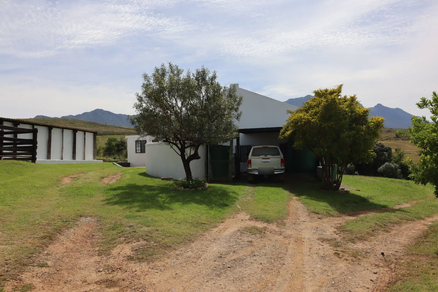 The farm road up to a white house under trees, with a carport and the mountains behind