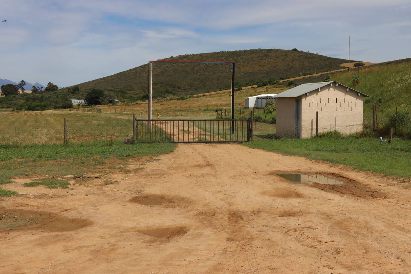 A farm gate on the sand road, with a small building beside it and a hill behind