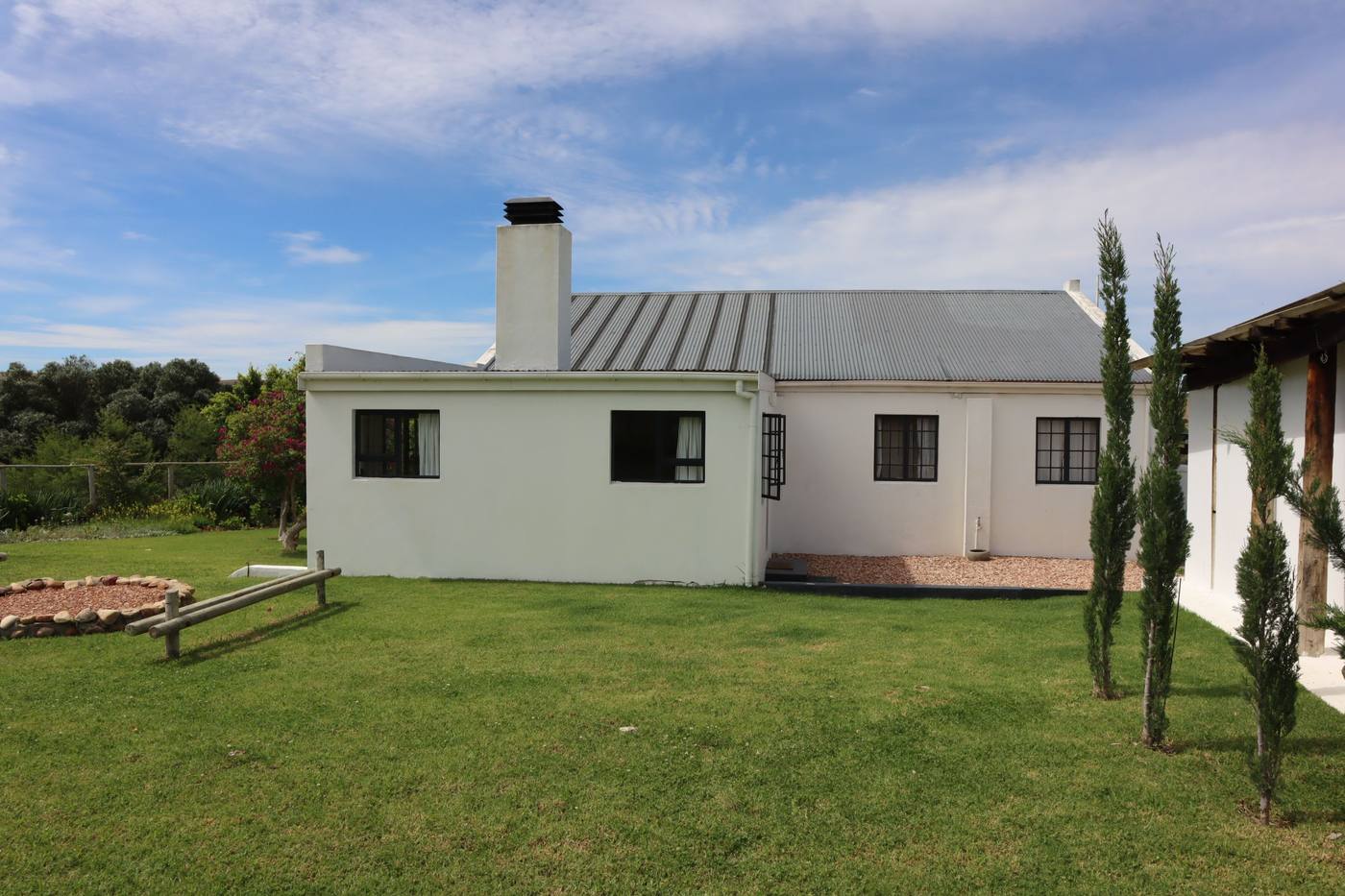 A white house with a chimney and a corrugated roof, with a fire pit and a bench on the lawn