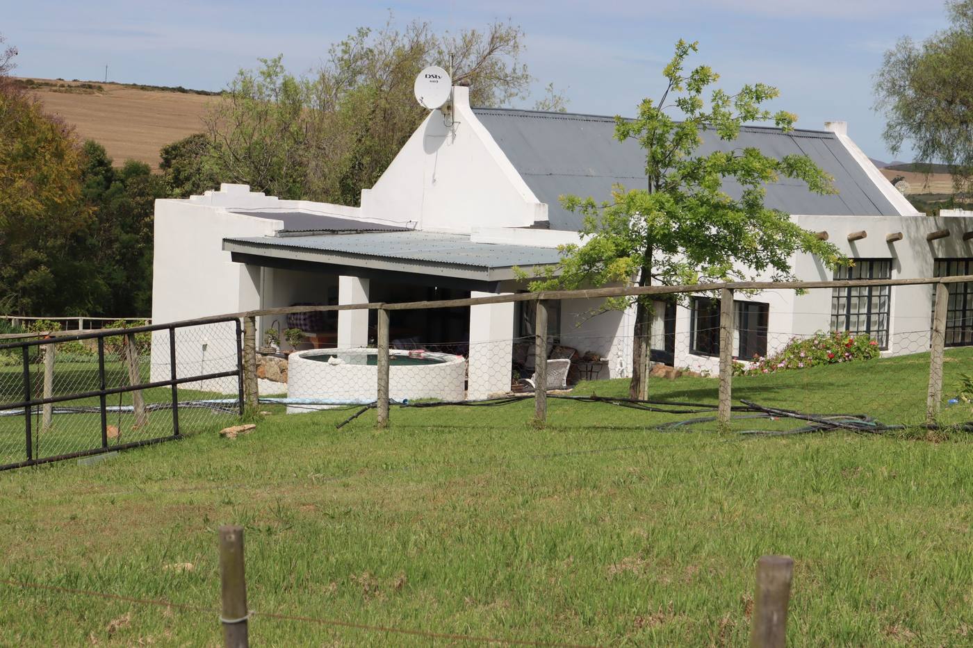 A white house with a gable and a satellite dish, behind a fenced lawn and a young tree